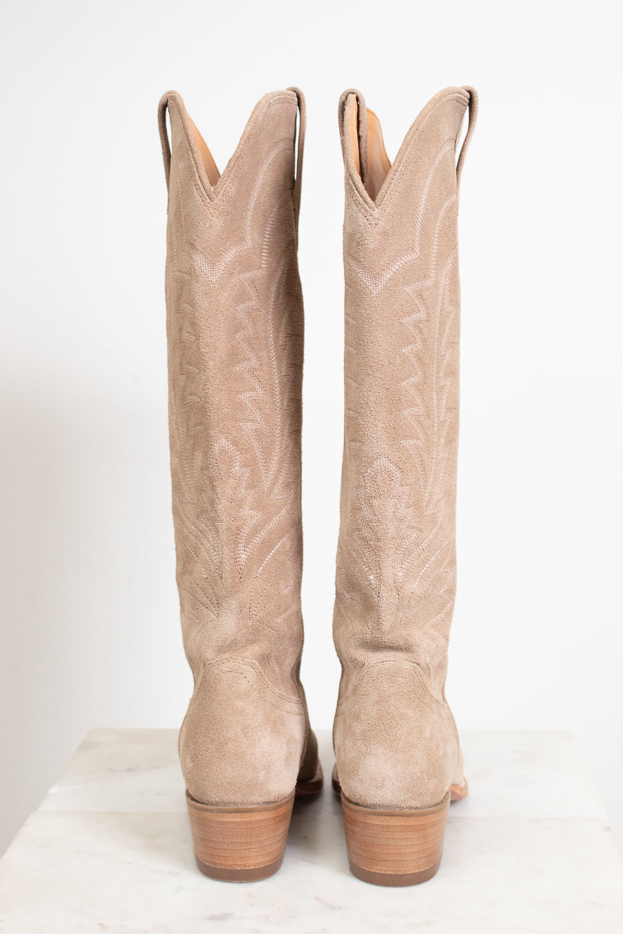 A pair of tall western-style boots in light tan suede, shown from the back on a white marble surface against a white background.