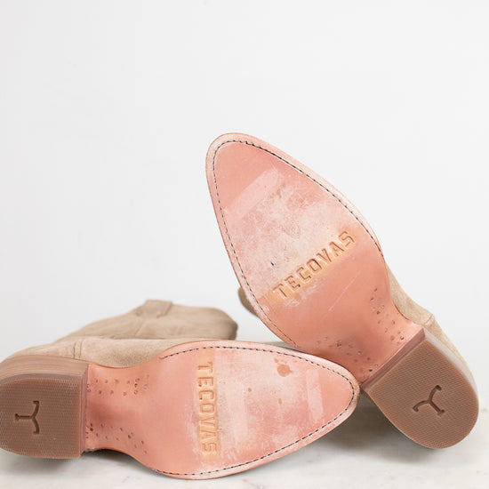 A pair of light tan suede boots turned over to show the insoles, which are a dusty pink color and embossed with the brand name 'TECOVAS'.