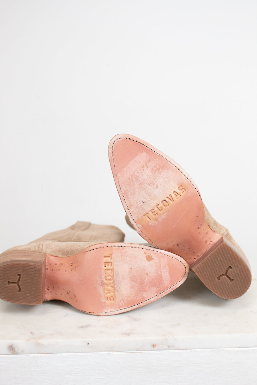 A pair of light tan suede boots turned over to show the insoles, which are a dusty pink color and embossed with the brand name 'TECOVAS'.