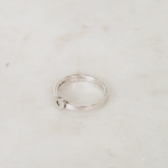 A thin silver ring with a small heart-shaped detail on the side, resting on a light beige marble surface.