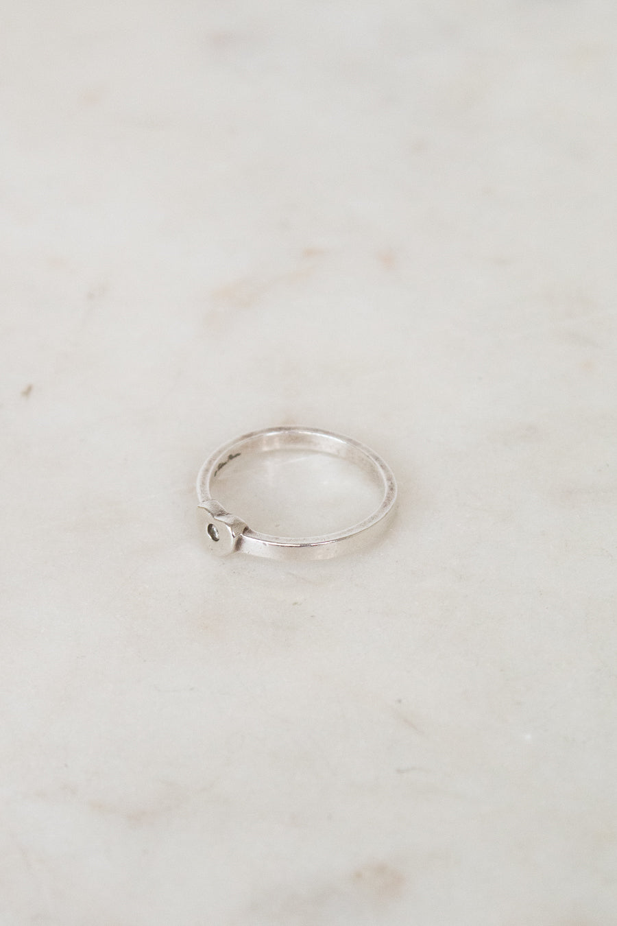 A thin silver ring with a small heart-shaped detail on the side, resting on a light beige marble surface.