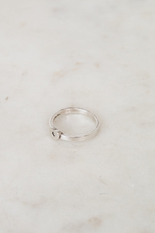 A thin silver ring with a small heart-shaped detail on the side, resting on a light beige marble surface.