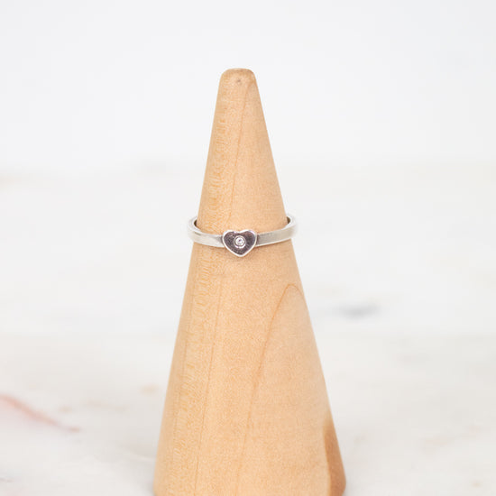 A silver ring with a small heart-shaped purple stone, displayed on a light wood cone stand against a white background.