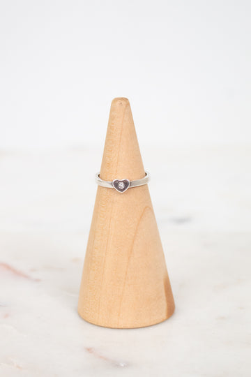 A silver ring with a small heart-shaped purple stone, displayed on a light wood cone stand against a white background.