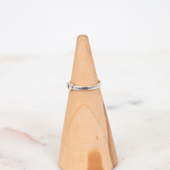 A thin silver band ring with a small clear gemstone, displayed on a light wood cone ring holder over a white marble surface.