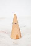 A thin silver band ring with a small clear gemstone, displayed on a light wood cone ring holder over a white marble surface.
