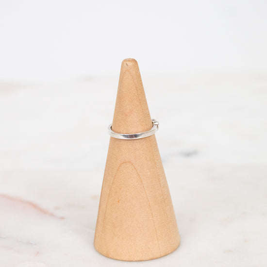 A thin silver band ring displayed on a light wood cone ring holder over a white marble surface.