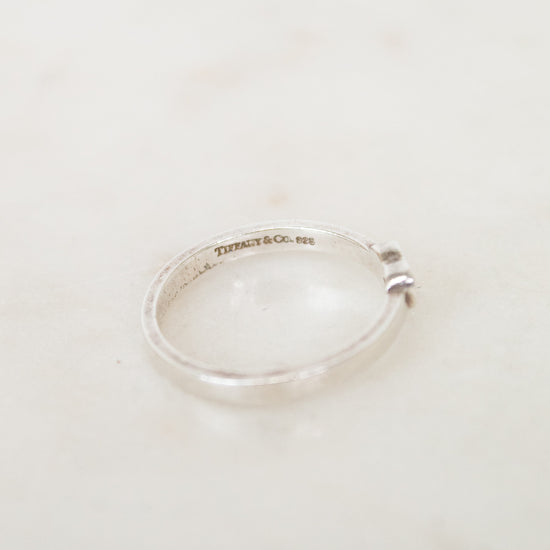 A silver open band ring with a small side accent, resting on a light beige marble surface. The inside of the band features a visible engraving reading 'Tiffany & Co.