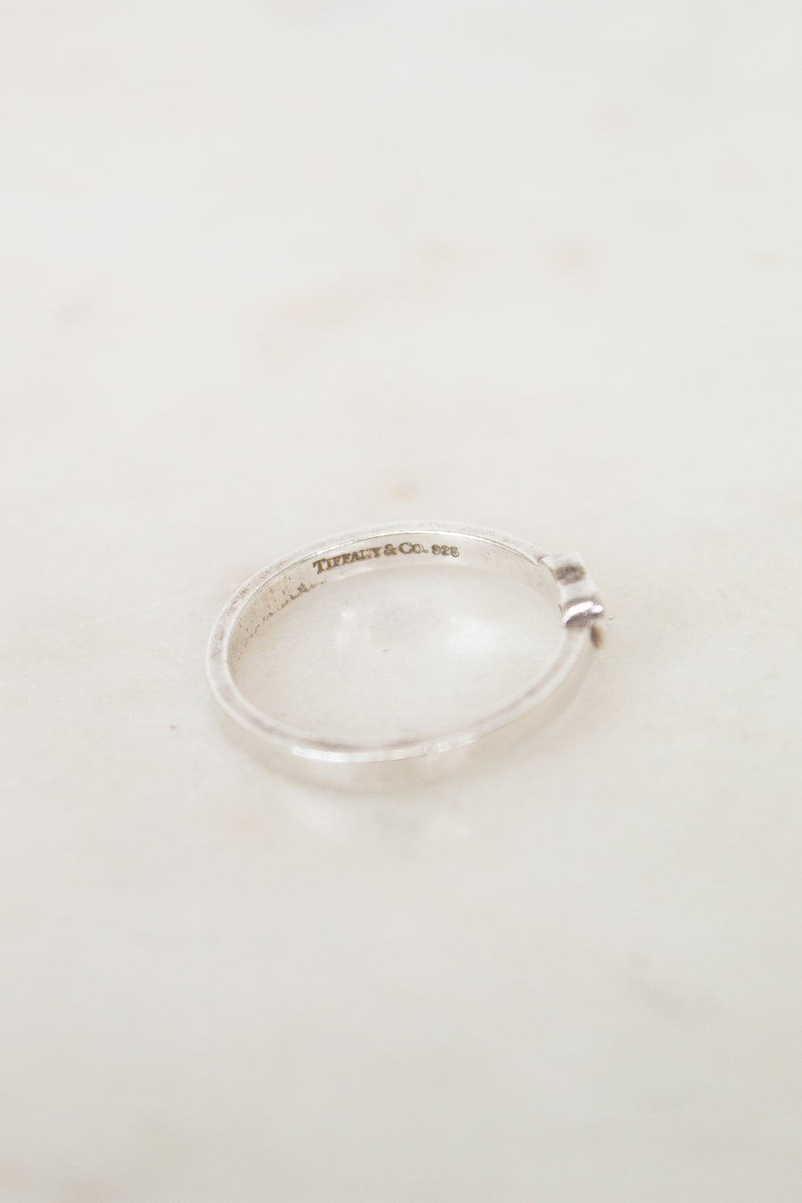 A silver open band ring with a small side accent, resting on a light beige marble surface. The inside of the band features a visible engraving reading 'Tiffany & Co.