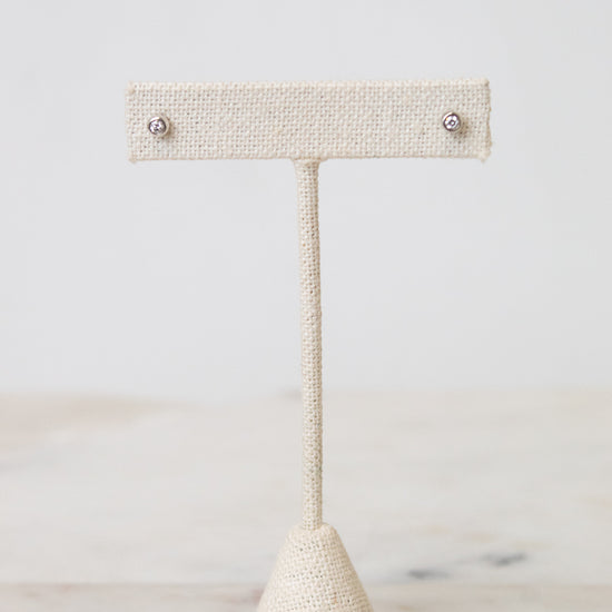 Front view of TIFFANY & CO. silver diamond circular stud earring resting on a beige earring stand , with a white and marble background.