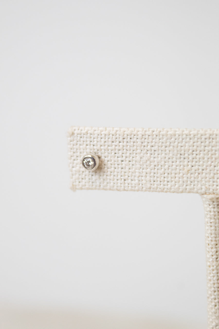 A close-up shot of one of the diamond studs, it is a circular shape with a diamond in the middle. The earring stud rest on a beige earring stand. 