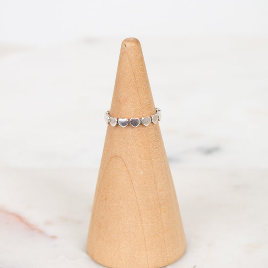 A delicate silver band ring with small heart charms, displayed on a light wood cone stand against a soft white marble background.