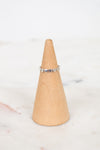 A delicate silver band ring with small heart charms, displayed on a light wood cone stand against a soft white marble background.