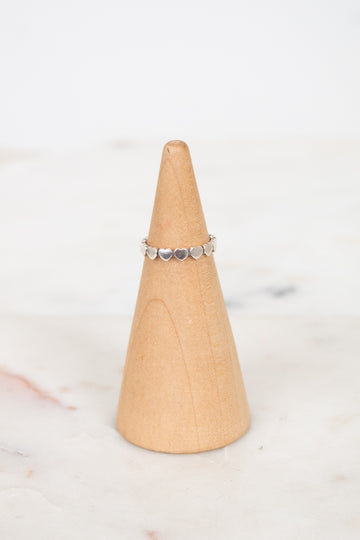 A delicate silver band ring with small heart charms, displayed on a light wood cone stand against a soft white marble background.