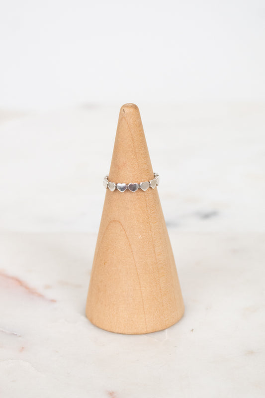 A delicate silver band ring with small heart charms, displayed on a light wood cone stand against a soft white marble background.