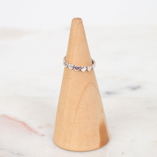 A delicate silver band ring with small heart-shaped clear stones, displayed on a light wood cone stand against a soft white marble background.