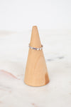 A delicate silver band ring with small heart-shaped clear stones, displayed on a light wood cone stand against a soft white marble background.