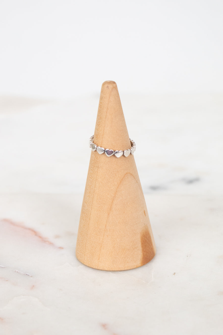 A delicate silver band ring with small heart-shaped clear stones, displayed on a light wood cone stand against a soft white marble background.
