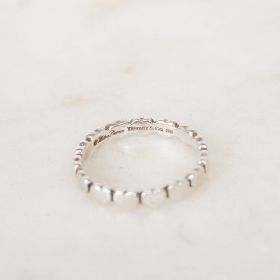 A delicate sterling silver beaded ring resting on a light beige marble surface. The inner band is engraved with 'TIFFANY & CO '.