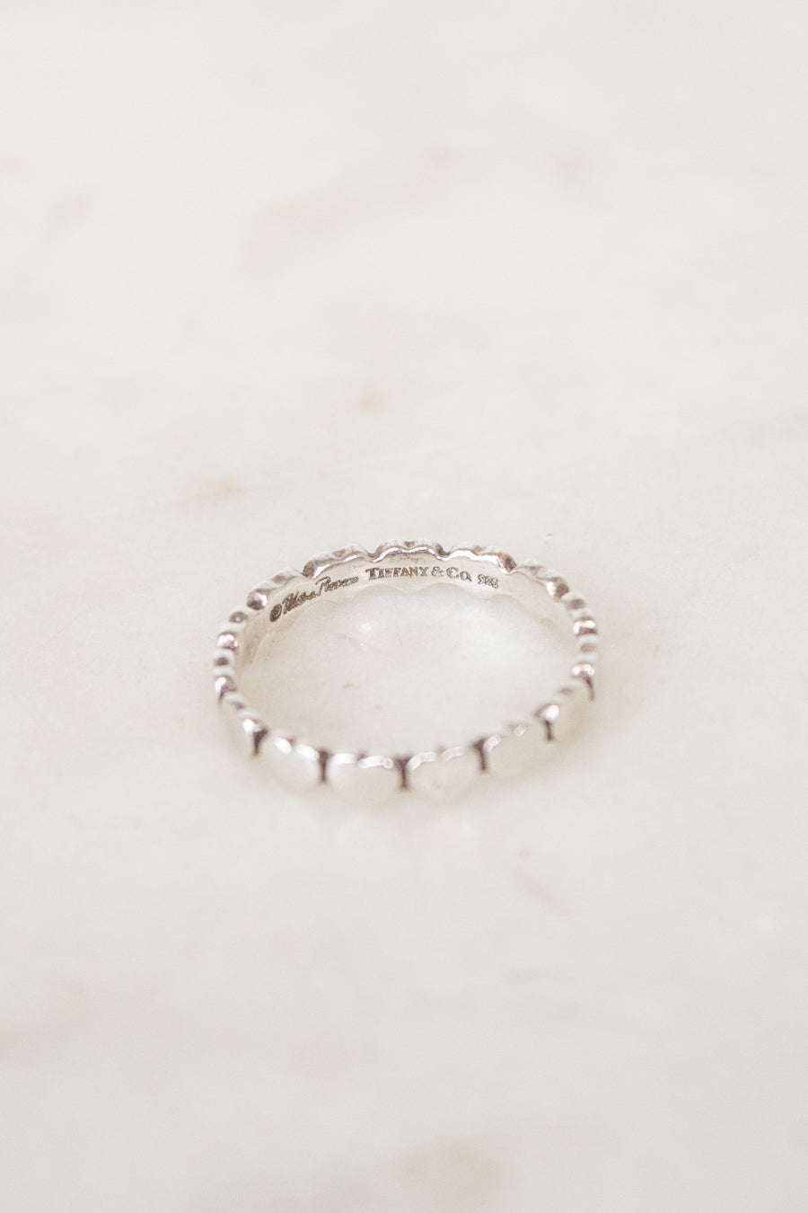 A delicate sterling silver beaded ring resting on a light beige marble surface. The inner band is engraved with 'TIFFANY & CO '.