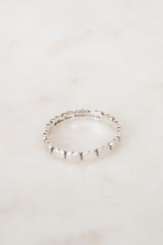 A delicate sterling silver beaded ring resting on a light beige marble surface. The inner band is engraved with 'TIFFANY & CO '.