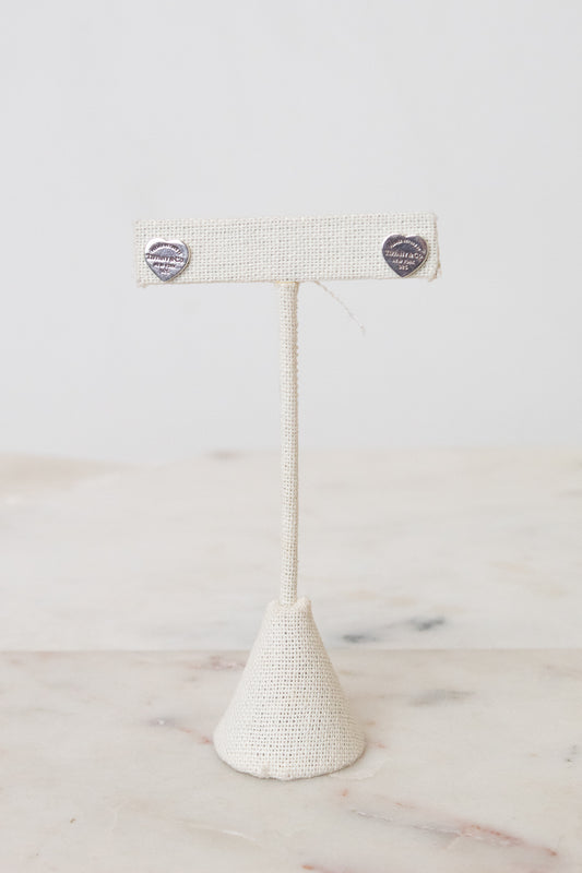 Pair of silver-tone Tiffany & Co , heart stud earring with engraved detailing, on a beige earring stand placed on a marble background.