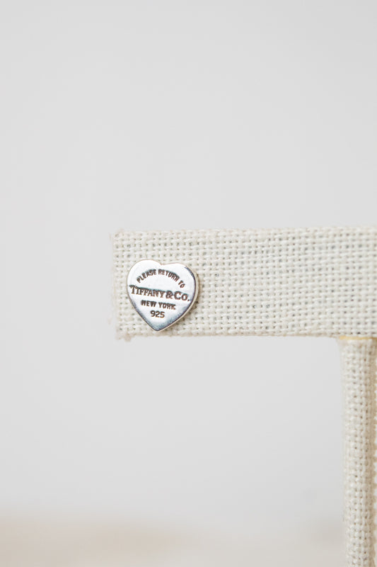 Close - up of a silver Tiffany & Co. heart-shaped charm engraved "PLEASE RETURN TO TIFFANY & CO, NEW YORK 925". The earrings are resting on a beige woven stand.