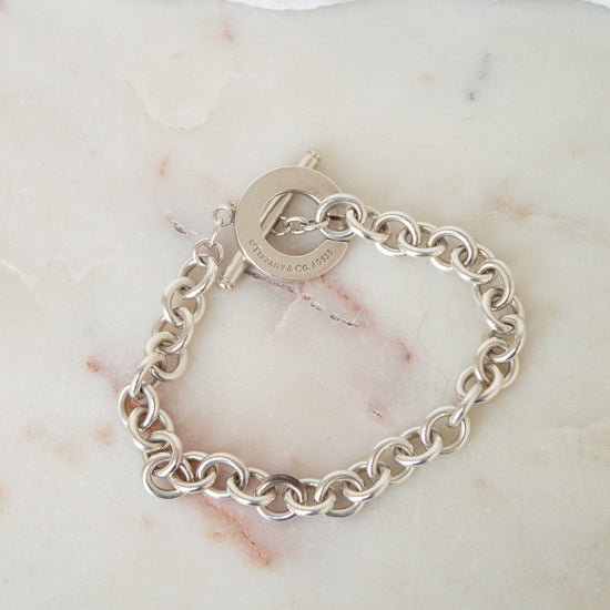 A silver chain link bracelet with a toggle clasp, sitting on a marble surface. 