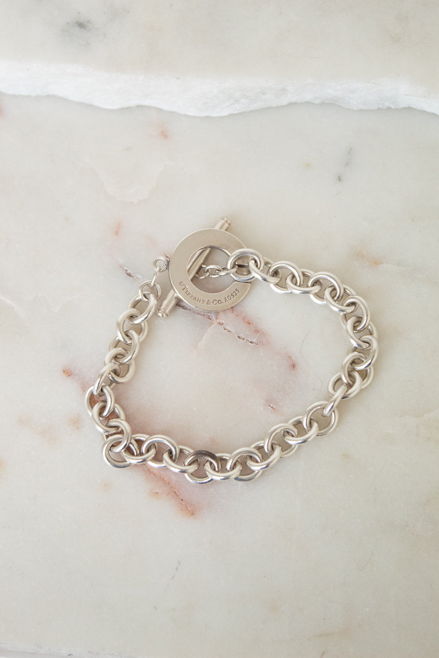 A silver chain link bracelet with a toggle clasp, sitting on a marble surface. 
