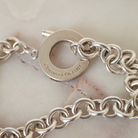 A close-up of a silver chain bracelet with a circular clasp engraved with 'TIFFANY & CO. ', resting on a light beige marble surface.