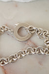 A close-up of a silver chain bracelet with a circular clasp engraved with 'TIFFANY & CO. ', resting on a light beige marble surface.
