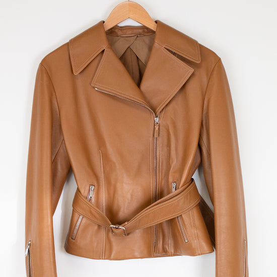 A tan leather moto jacket with a matching waist belt, hanging on a wooden hanger against a plain white wall.