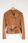 A tan leather moto jacket with a matching waist belt, hanging on a wooden hanger against a plain white wall.