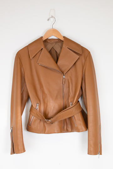 A tan leather moto jacket with a matching waist belt, hanging on a wooden hanger against a plain white wall.