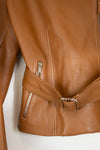 Close-up of a tan leather jacket showing the side zip pocket, belt detail with silver buckle, and zipper on a plain white background.
