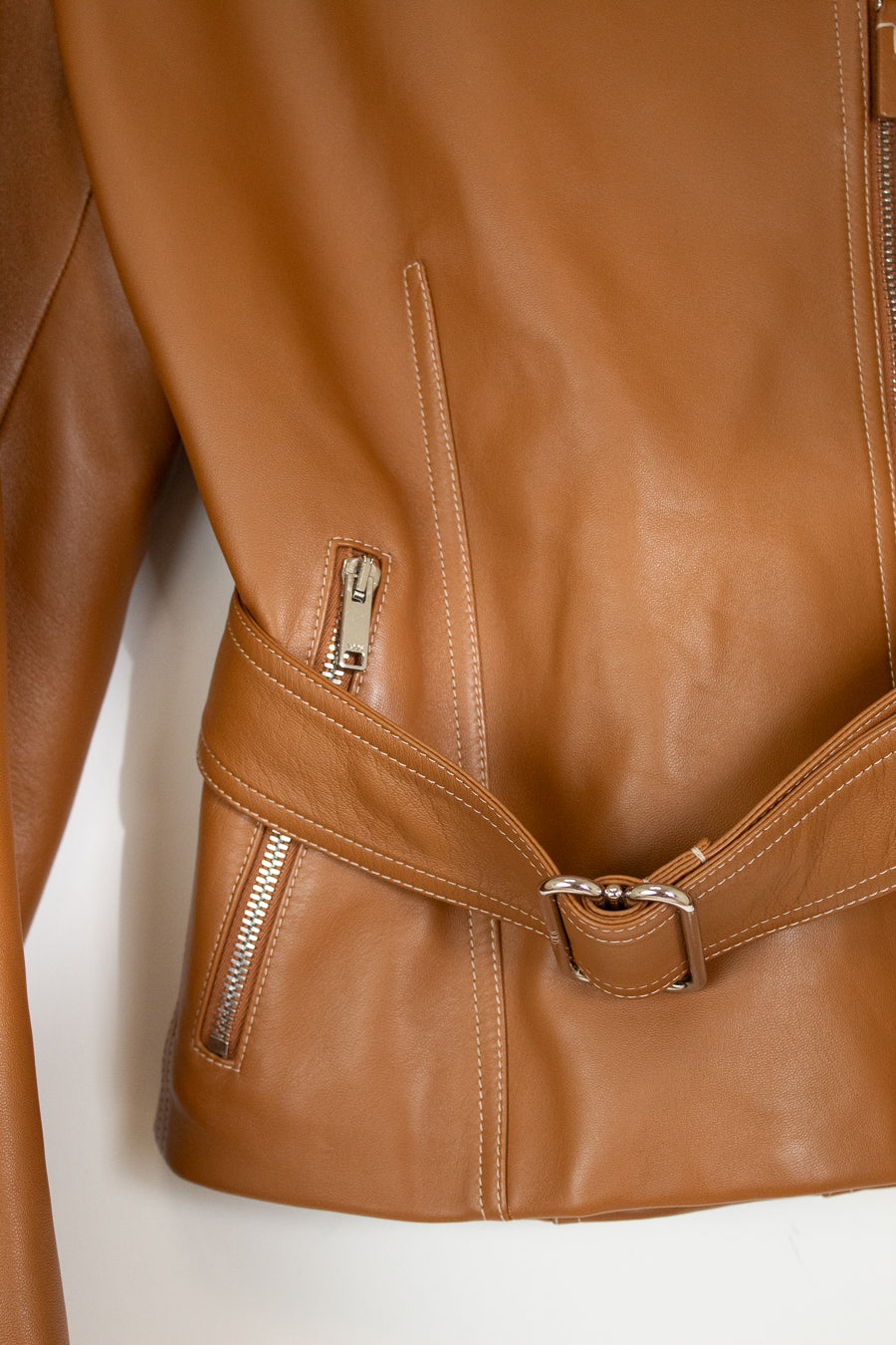 Close-up of a tan leather jacket showing the side zip pocket, belt detail with silver buckle, and zipper on a plain white background.