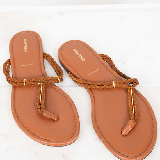 A pair of tan leather flat thong sandals with braided rope straps and gold-tone metal accents, displayed on a white marble surface.