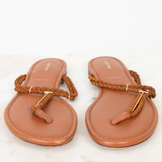 A pair of tan leather thong sandals with braided brown straps and gold-tone hardware, displayed on a white marble surface against a white background.