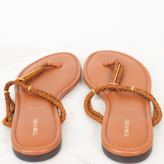 A pair of TOM FORD flat thong sandals in tan leather with braided brown straps and gold-tone hardware, shown from a top-down view on a white marble surface.