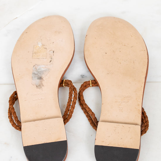 A pair of flat thong sandals shown from the bottom, featuring tan leather soles with a black toe cap and brown braided straps, laid flat on a white marble surface.