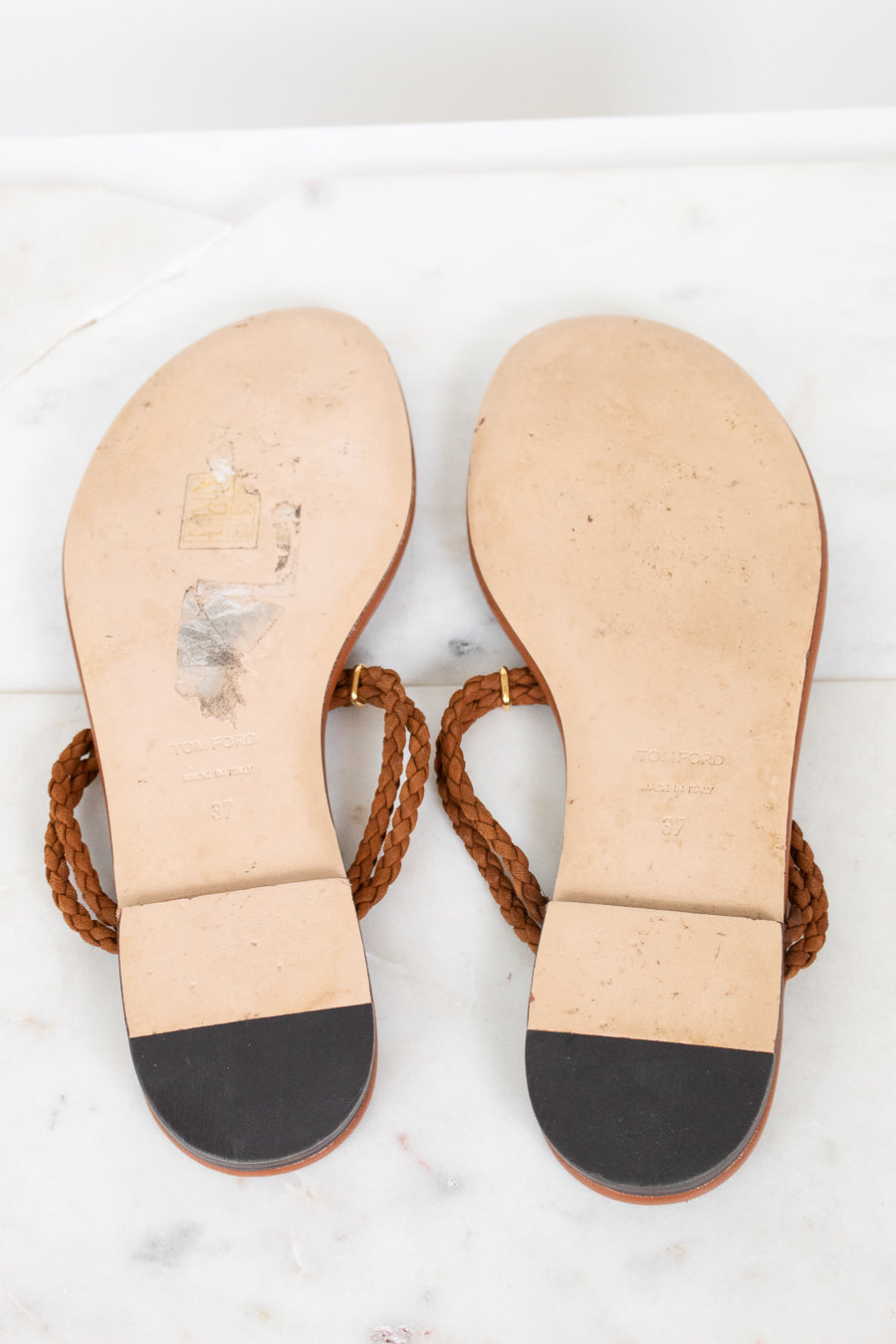 A pair of flat thong sandals shown from the bottom, featuring tan leather soles with a black toe cap and brown braided straps, laid flat on a white marble surface.