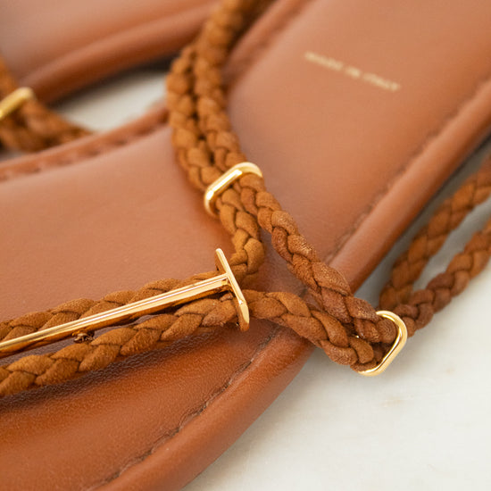 Close-up of a tan leather flat sandal with braided brown straps and gold-tone metal accents, resting on a light surface.