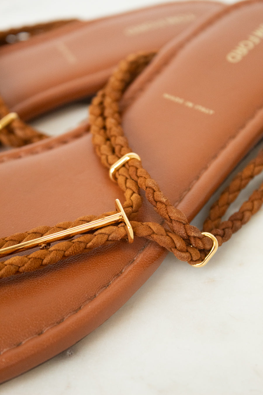 Close-up of a tan leather flat sandal with braided brown straps and gold-tone metal accents, resting on a light surface.