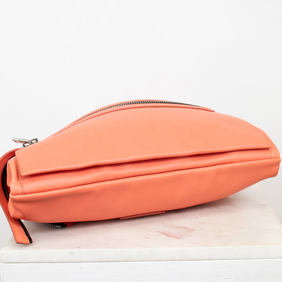 A coral orange leather crossbody bag with a top zipper, resting on a white marble block against a white background.