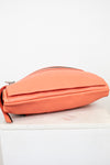 A coral orange leather crossbody bag with a top zipper, resting on a white marble block against a white background.