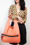 A woman wearing a tan and black polka-dot blouse and black skirt holds a large coral-orange shoulder bag with black zipper details against a plain white background.