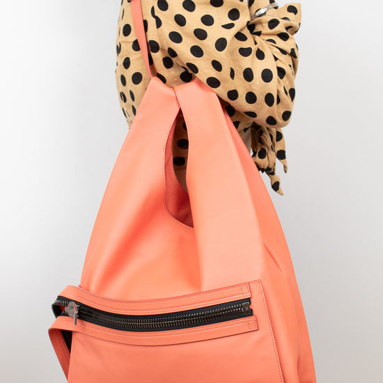 A woman wearing a beige and black polka-dot blouse carries a large, structured coral-orange tote bag with a black zippered pocket on the front, set against a plain light gray background.