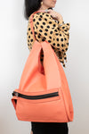 A woman wearing a beige and black polka-dot blouse carries a large, structured coral-orange tote bag with a black zippered pocket on the front, set against a plain light gray background.