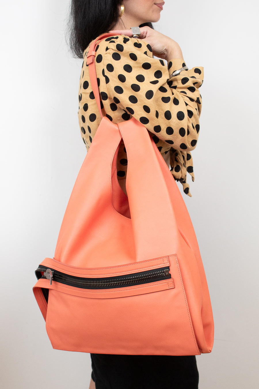A woman wearing a beige and black polka-dot blouse carries a large, structured coral-orange tote bag with a black zippered pocket on the front, set against a plain light gray background.