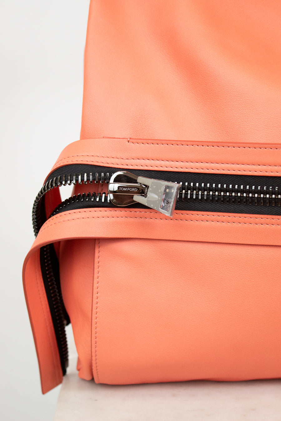 Close-up of a coral orange leather bag with a black zipper and a silver-toned metal slider pull featuring the 'Tory Ford' brand name.